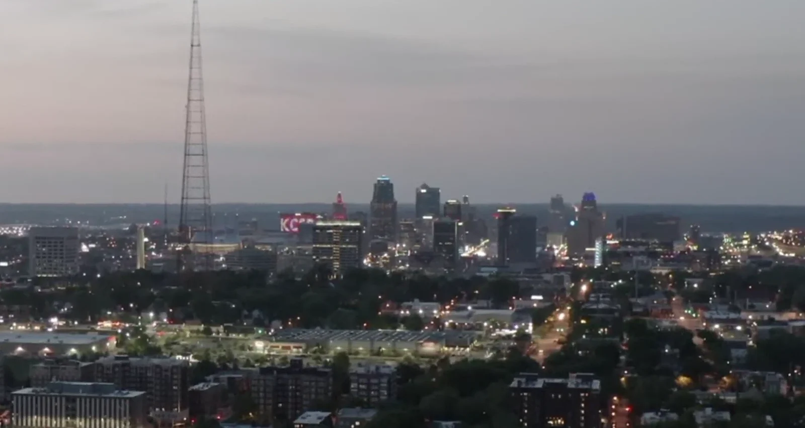 The downtown Kansas City skyline at dusk, photographed from the air, with the broadcast tower standing in the foreground and the lit KC sign below it.